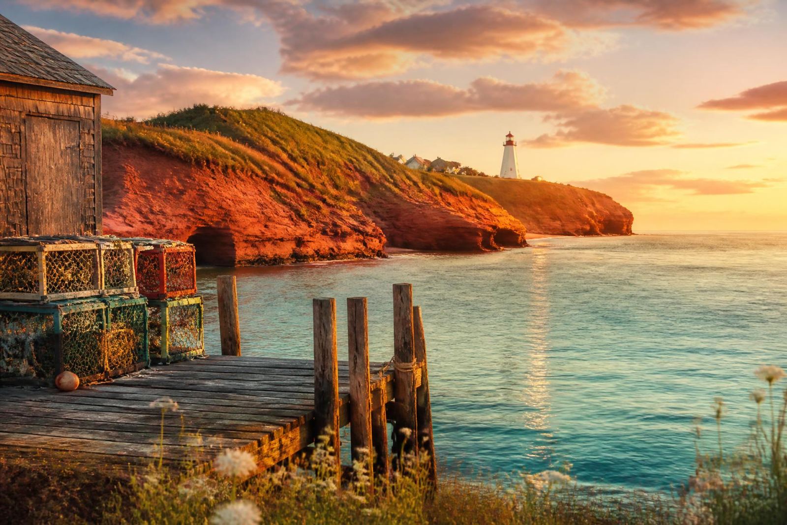 Red sandstone cliffs, a wooden wharf with lobster traps, and a lighthouse at golden hour on Prince Edward Island
